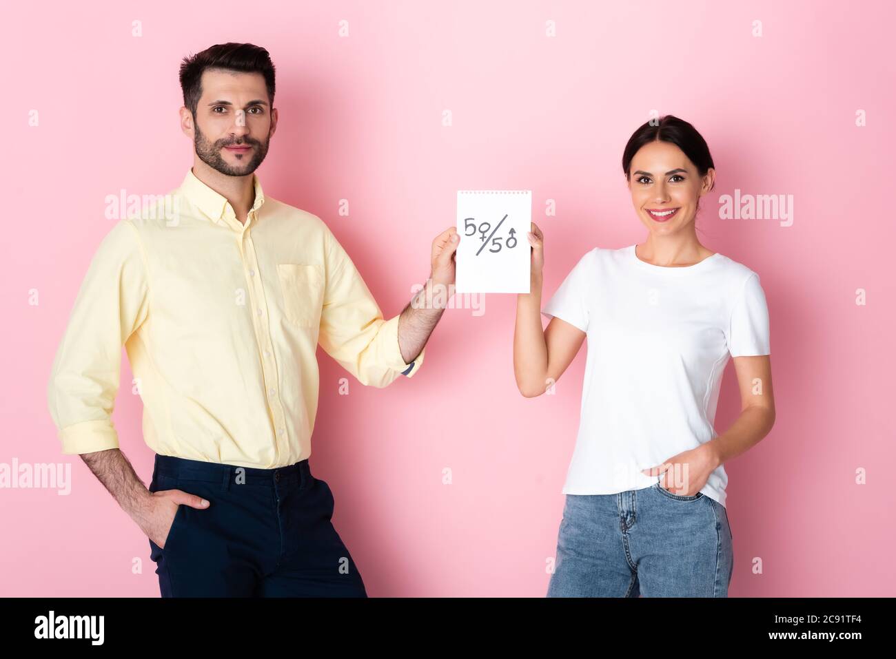 happy man and woman en t-shirt blanc tenant du papier avec cinquante symboles tout en se tenant avec les mains dans les poches sur le rose Banque D'Images