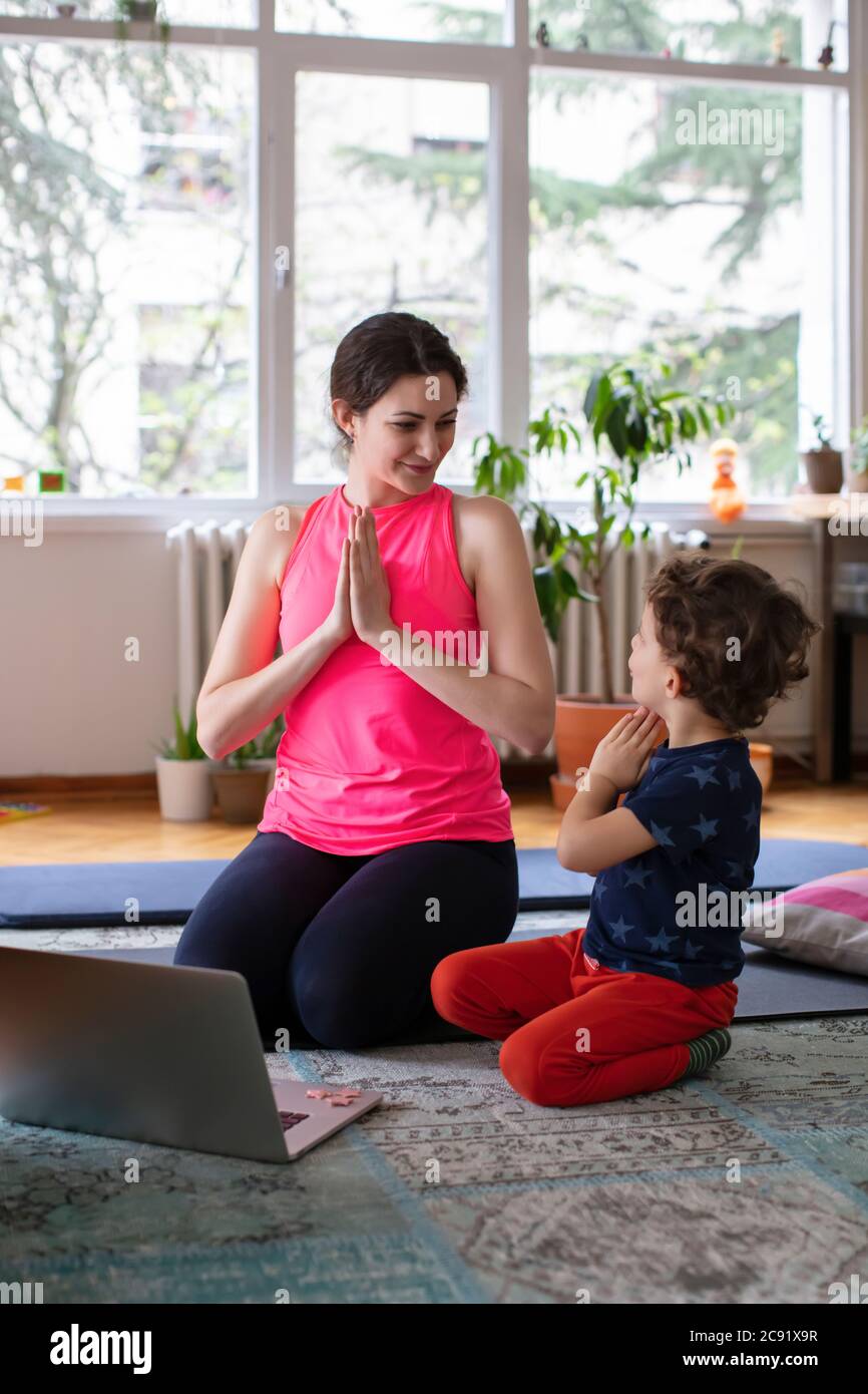 Jeune mère et enfant faisant Namaste poser tout en pratiquant le yoga À la maison, devant l'ordinateur portable Banque D'Images