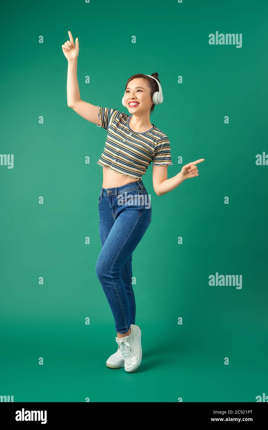 Portrait complet de la joyeuse femme asiatique sur des vêtements décontractés dansant et écoutant de la musique avec un casque sur fond vert. Banque D'Images