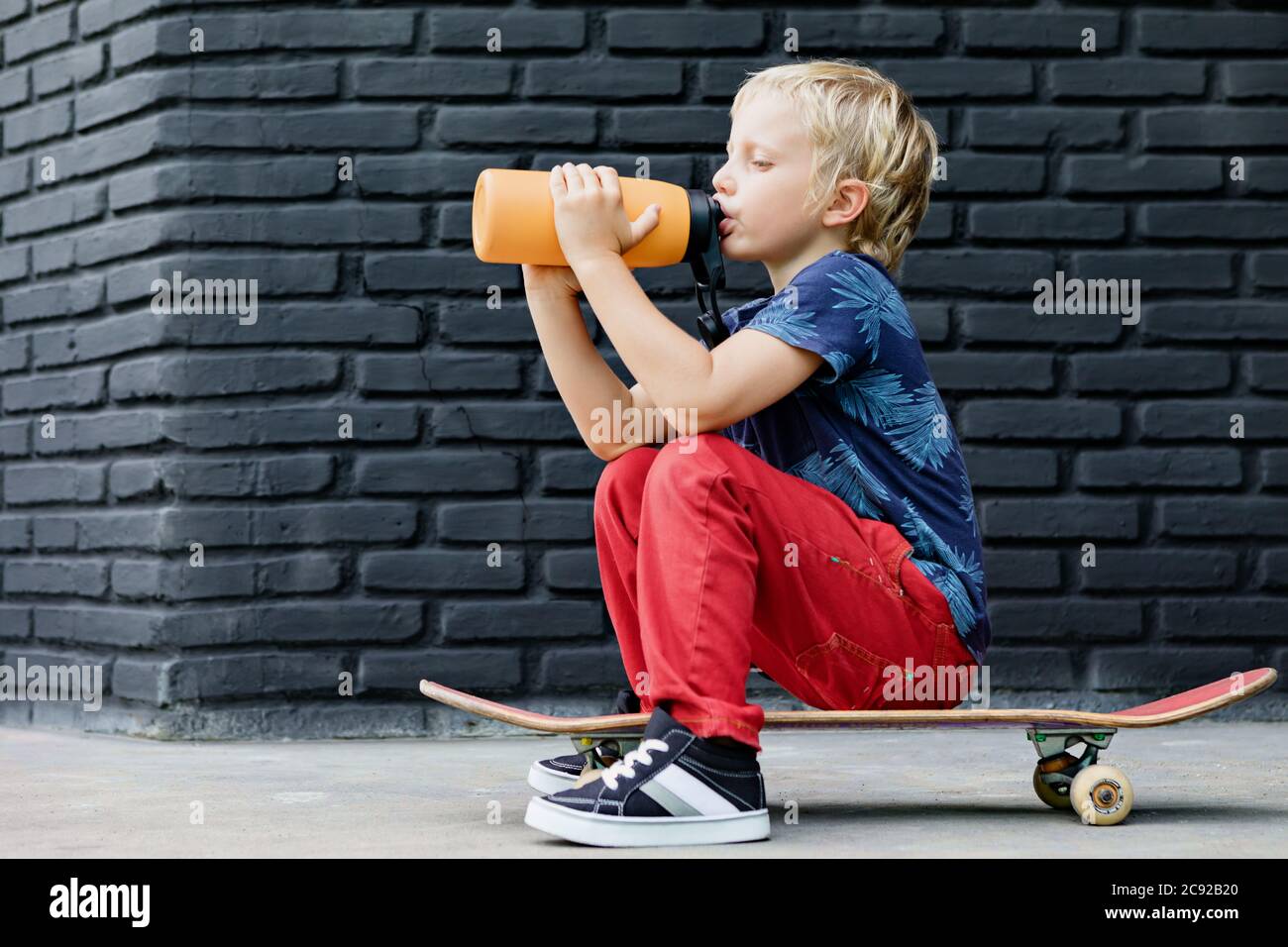 Le jeune patineur s'assoit sur le skateboard, boit de l'eau fraîche du biberon réutilisable après la formation des enfants. Vie familiale active, activités de plein air Banque D'Images