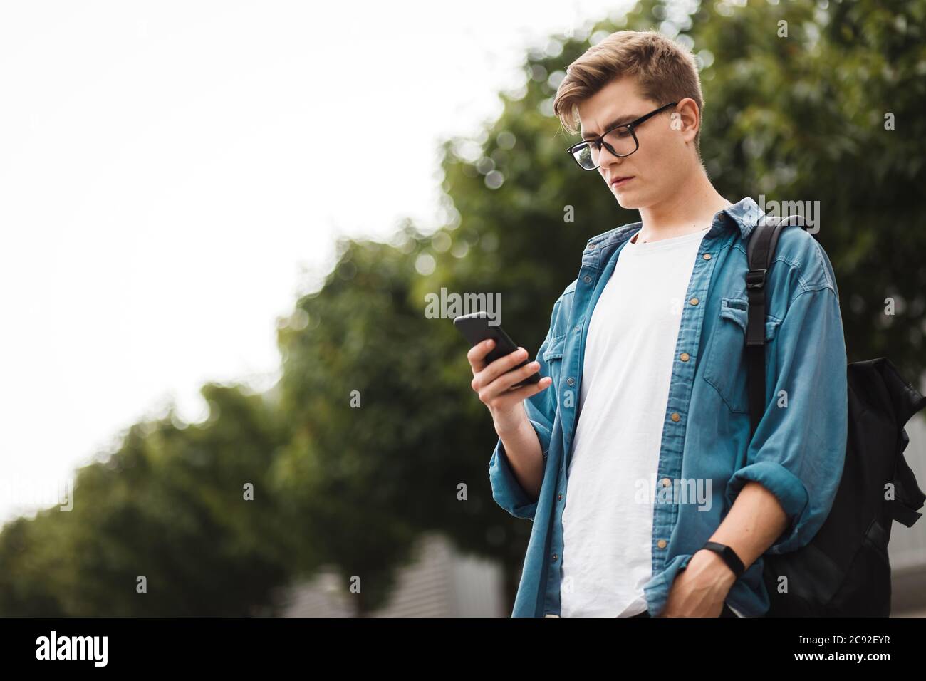 Jeune étudiant moderne de sexe masculin discutant sur le réseau en se tenant au milieu de la rue. Beau gars en lunettes avec un téléphone dans ses mains Banque D'Images