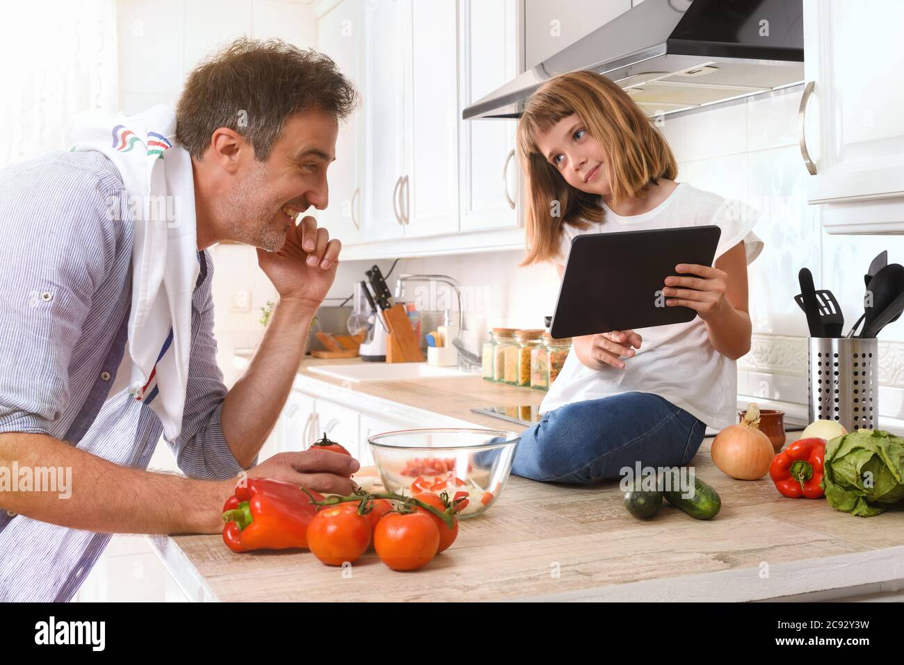 Fille assise sur un banc de cuisine montrant à son père une recette numérique à partir d'une tablette pour préparer un plat Banque D'Images