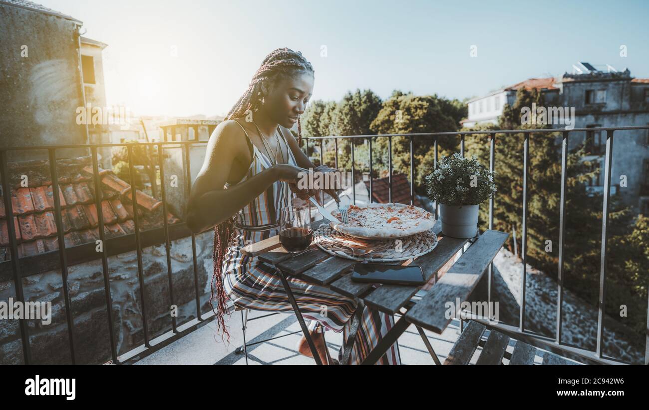 Une jeune femme noire ravishing prend le déjeuner et coupe une délicieuse pizza tout en étant assise à table avec un verre de vin Banque D'Images