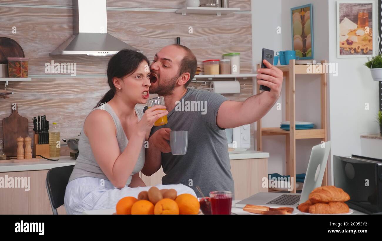 Un jeune couple heureux prend des selfies au petit déjeuner. Joyeux mari et femme mariés faisant des visages drôles tout en prenant une photo pendant le petit déjeuner dans la cuisine. Banque D'Images