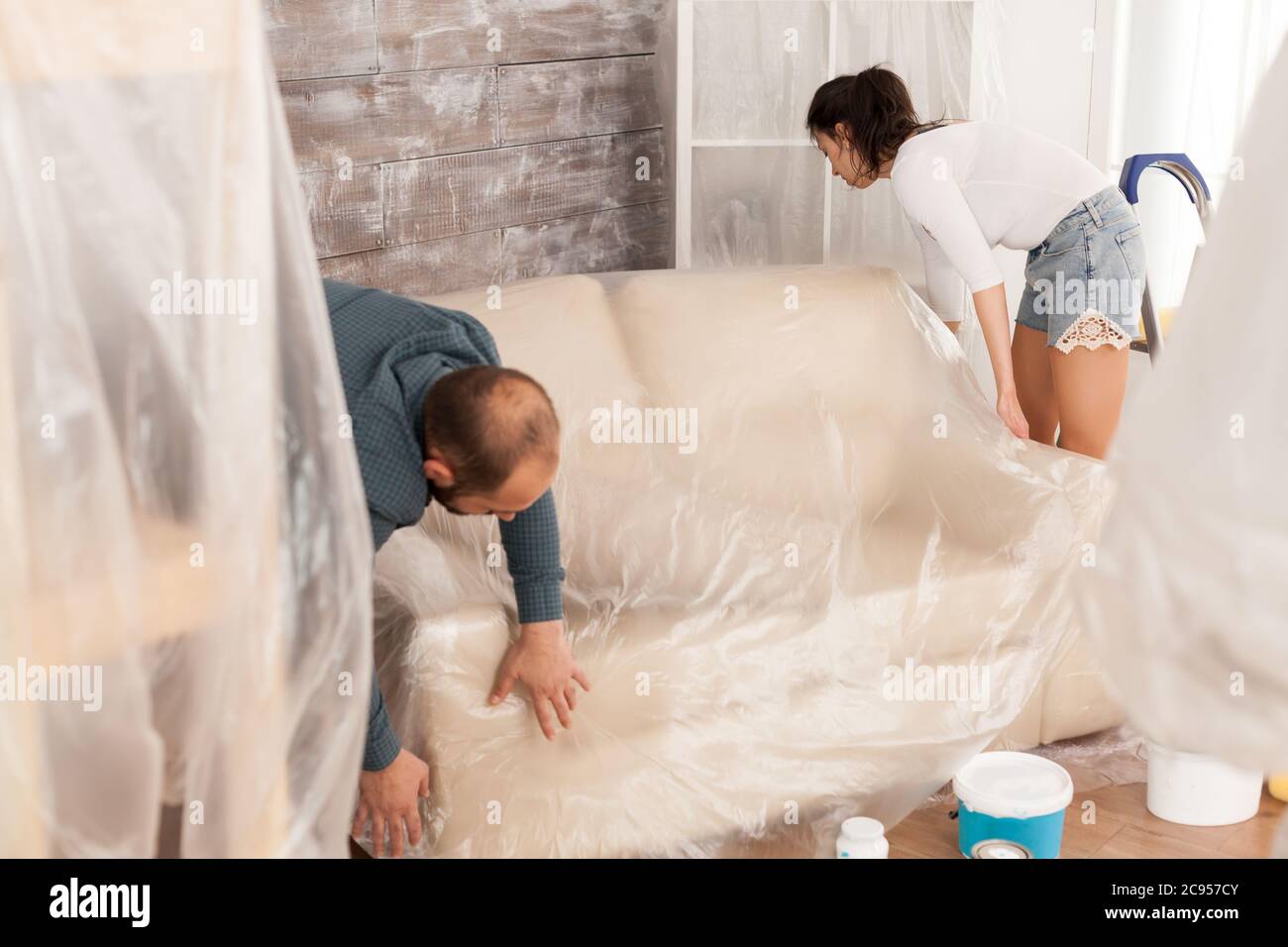 Un couple emballant le canapé dans un film plastique pour la protection  pendant qu'ils sont en train de rénover la salle de séjour. Maison pendant  la rénovation, la décoration et la peinture.