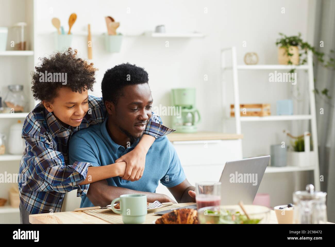 Homme africain assis à la table et travaillant sur ordinateur portable avec son fils qui l'embrasse, ils sont dans la cuisine Banque D'Images