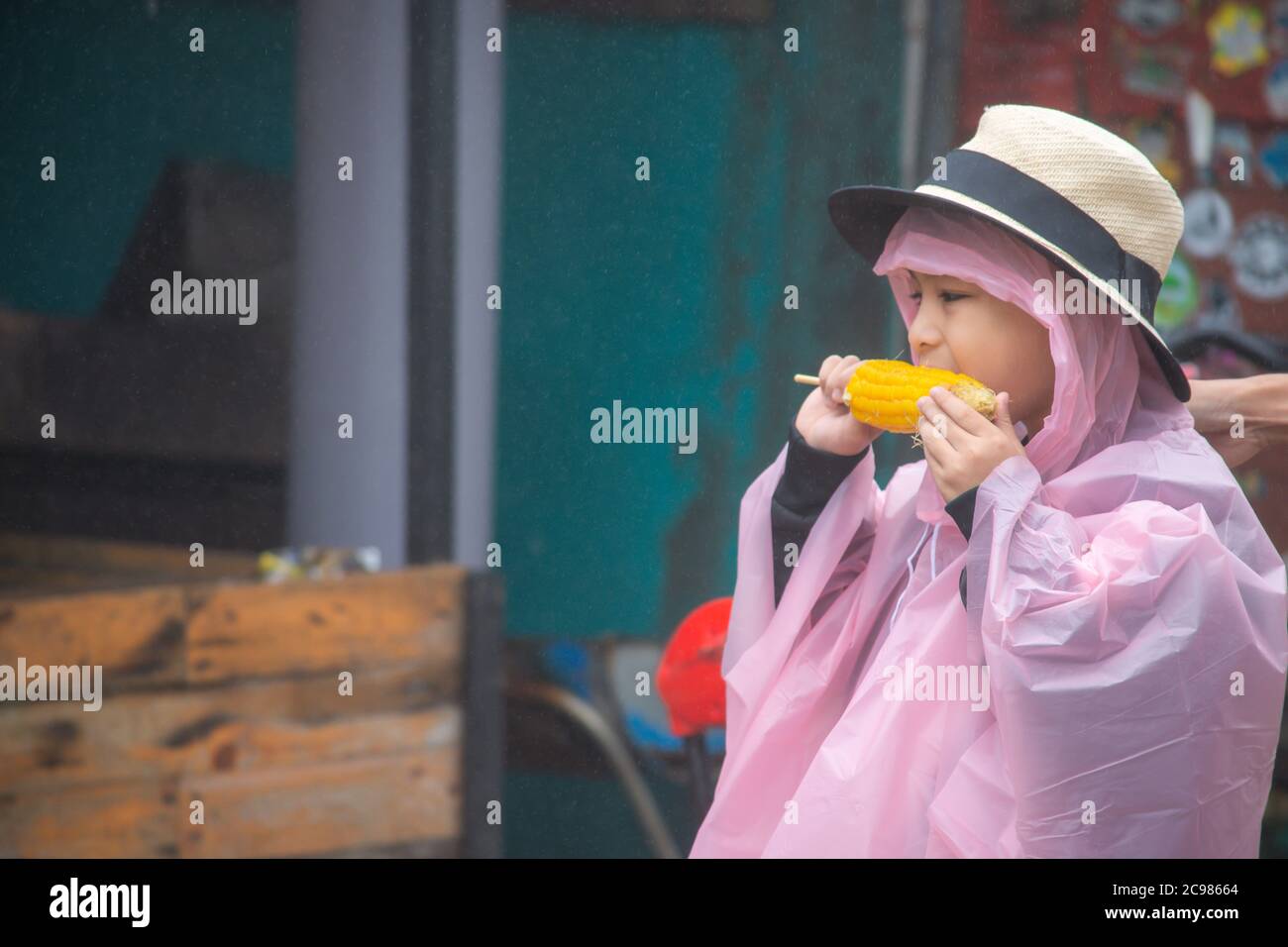 Garçon portant des vêtements de pluie Eating maïs dans la main et gouttelettes de pluie flottant dans l'air. Banque D'Images