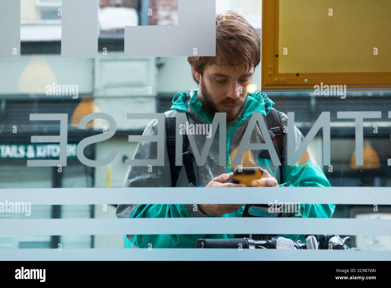 Deliveroo emporter repas courrier livraison cycliste à l'extérieur d'un restaurant d'où il fait une collection. Il vérifie son appareil mobile. Angleterre Royaume-Uni (120) Banque D'Images