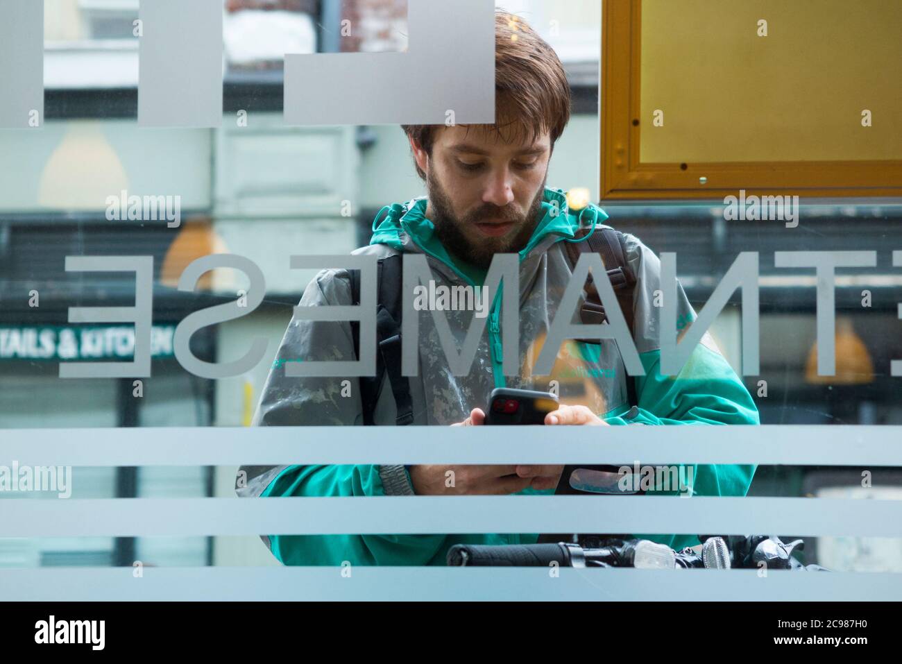 Deliveroo emporter repas courrier livraison cycliste à l'extérieur d'un restaurant d'où il fait une collection. Il vérifie son appareil mobile. Angleterre Royaume-Uni (120) Banque D'Images