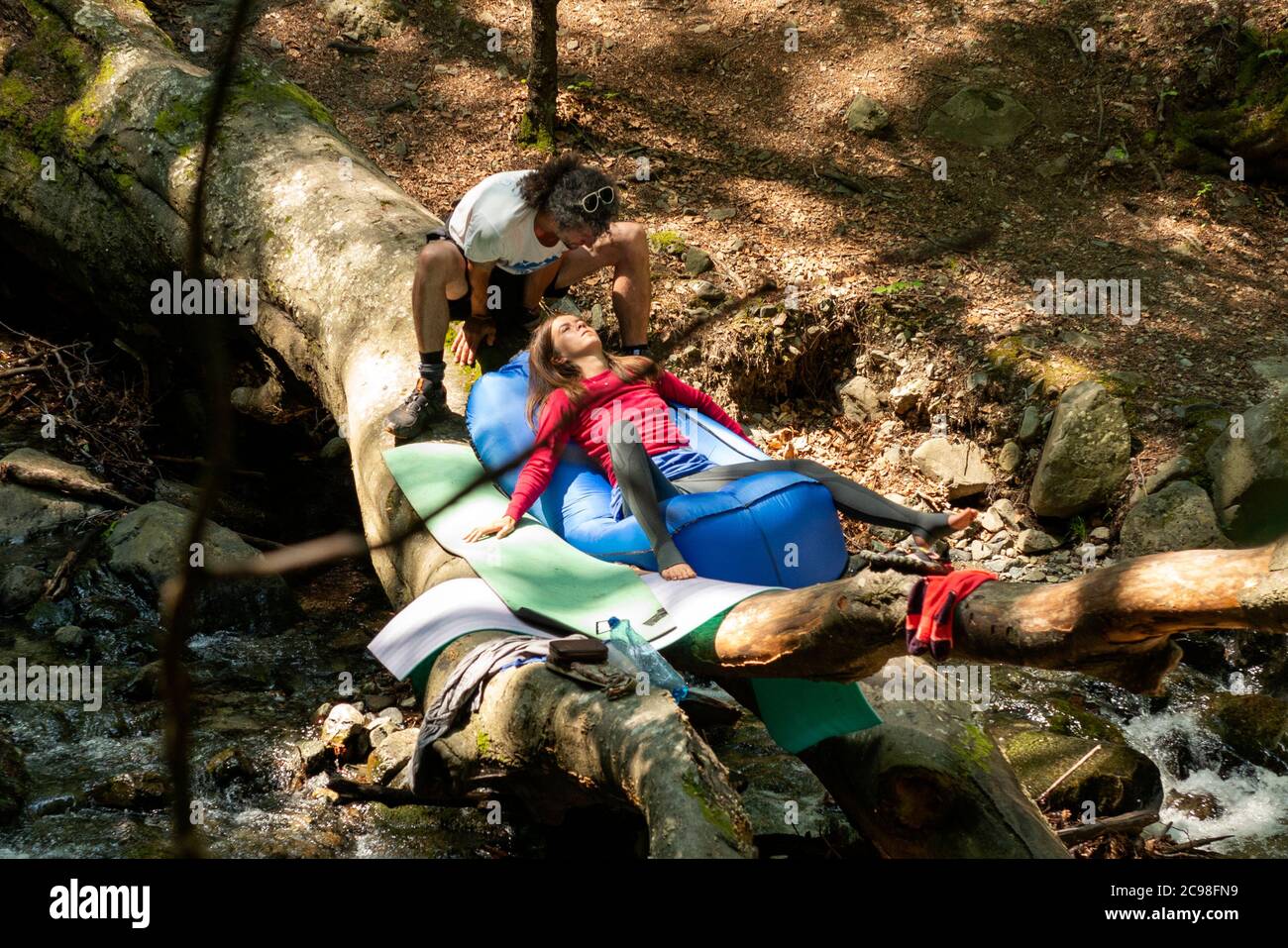 Couple de randonneurs se détendant sur un arbre déchu en bois au-dessus de la rivière montagne ruisseau en été ensoleillé comme la baignade de la forêt de yoku shinrin et l'évasion de la nature. Banque D'Images