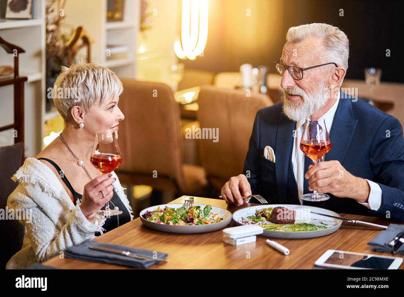 Un couple senior élégant et élégant se regarde les uns les autres et discute, entretient des relations étroites. Un homme aux cheveux gris en tuxedo lève un verre. Utilisez une cigarette électronique, un singe. Mo Banque D'Images