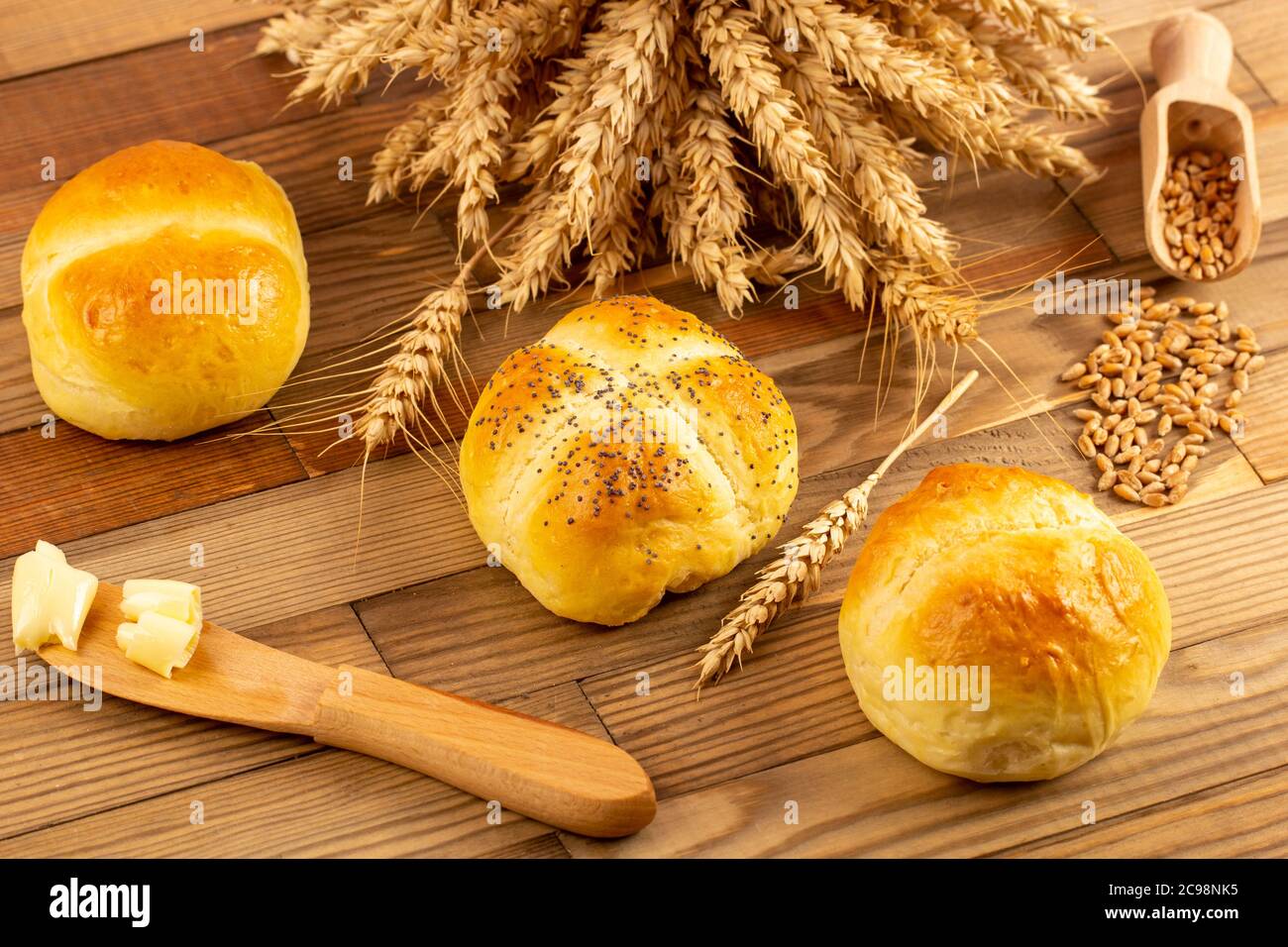 Trois petits pains posés sur une table en bois à côté des céréales et des épis de blé. À côté, il y a un couteau en bois avec du beurre. Banque D'Images