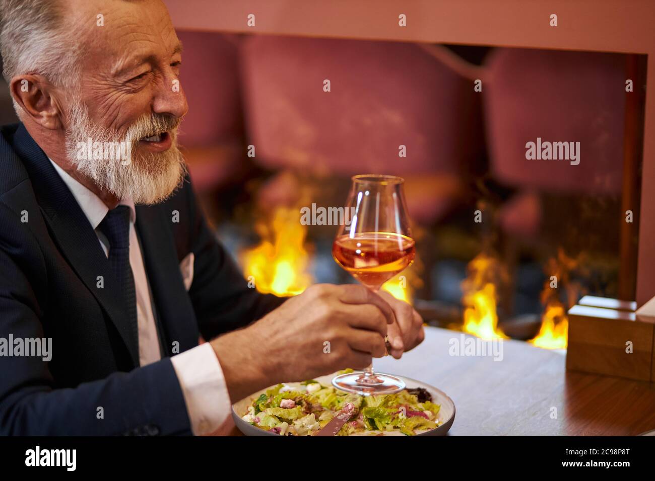 Homme à la barbe et aux cheveux gris en smoking avec verre de champagne, assis au restaurant et rire, regardez la personne en face. Contexte firemplacer Banque D'Images