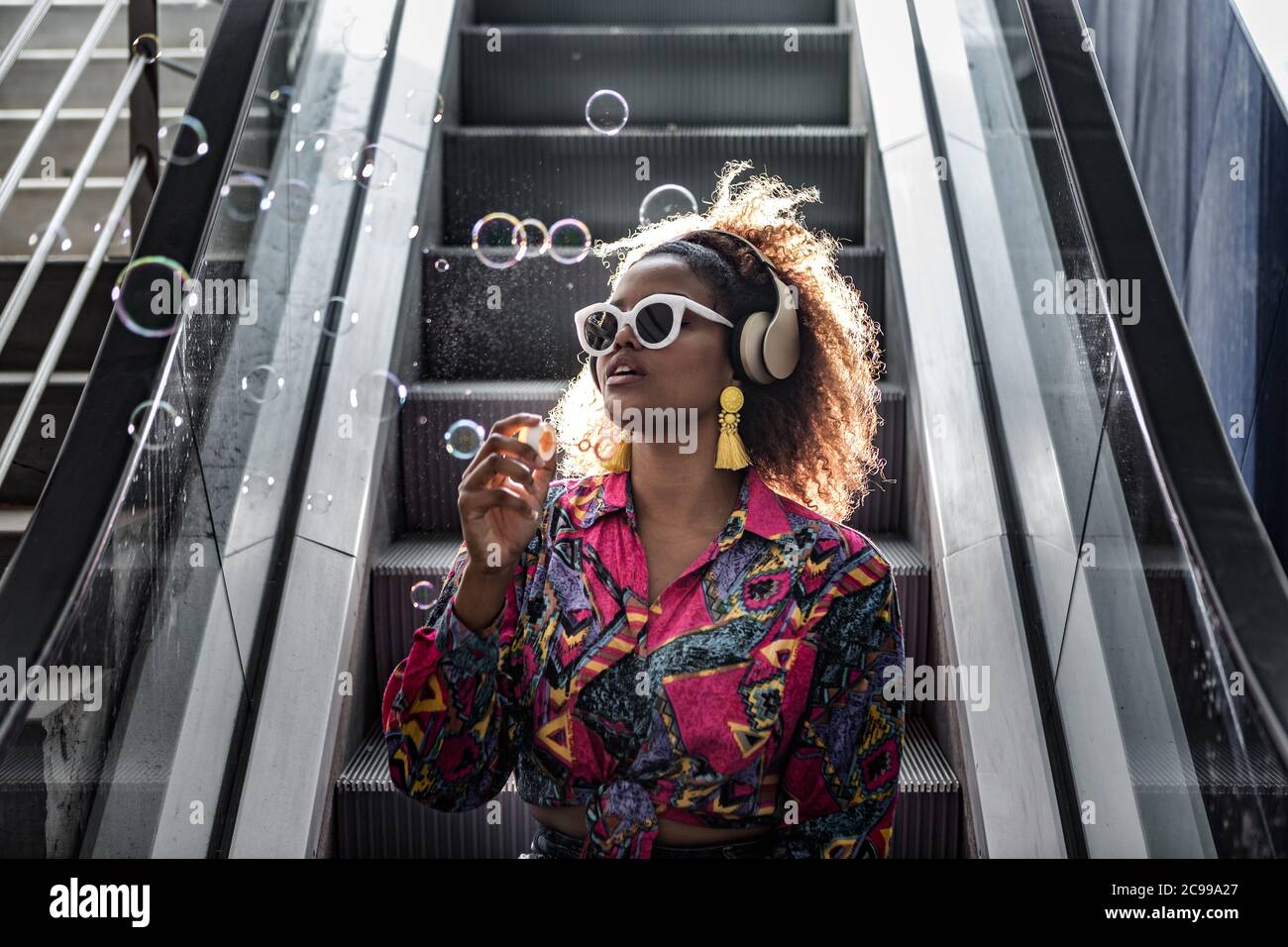 Femme afro-américaine en lunettes de soleil et blouse colorée assise sur un  escalier en mouvement avec une bouche ouverte tout en soufflant des bulles  dans le dos éclairé Photo Stock - Alamy