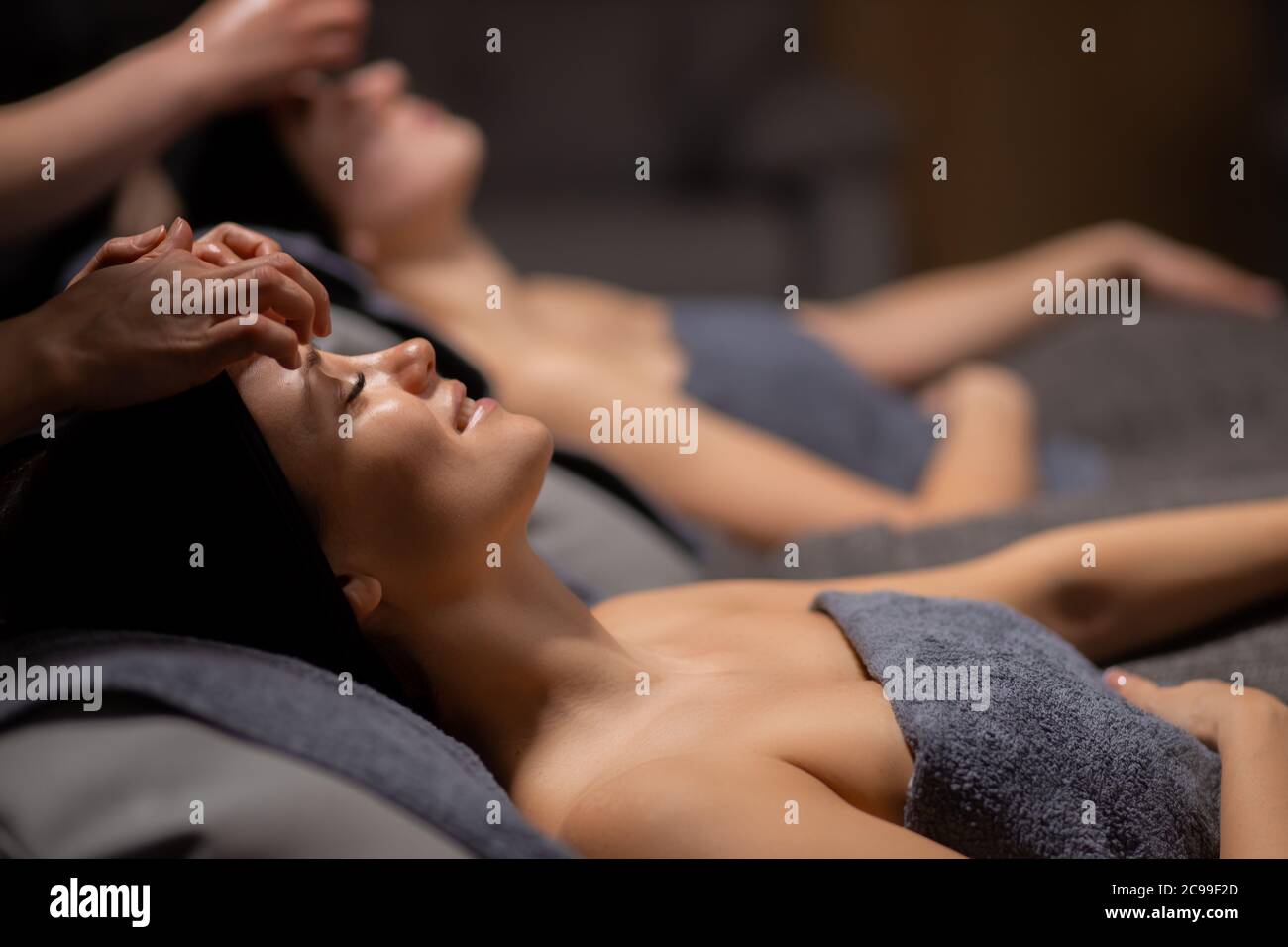 deux belles femmes caucasiennes reçoivent un massage facial, les jeunes femmes s'étendent sur le lit tandis que les masseurs touchent la peau de la tête et du visage, font le massage avec soin Banque D'Images