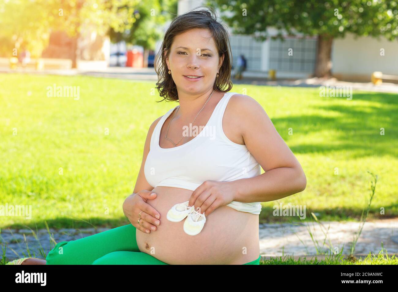 Femme Enceinte Touchant Son Ventre Ventre Ventre De Jouer Avec De Petites Chaussures De Bebe Assis A L Exterieur Sur Une Prairie D Herbe Verte Dans Un Parc De La Ville Par Une Journee