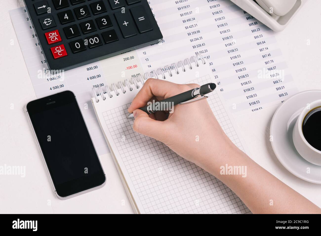 La main d'une femme écrit dans un carnet avec un crayon. Bureau avec une calculatrice, un téléphone portable, une tasse à café et des documents financiers. Concept de comptabilité Banque D'Images