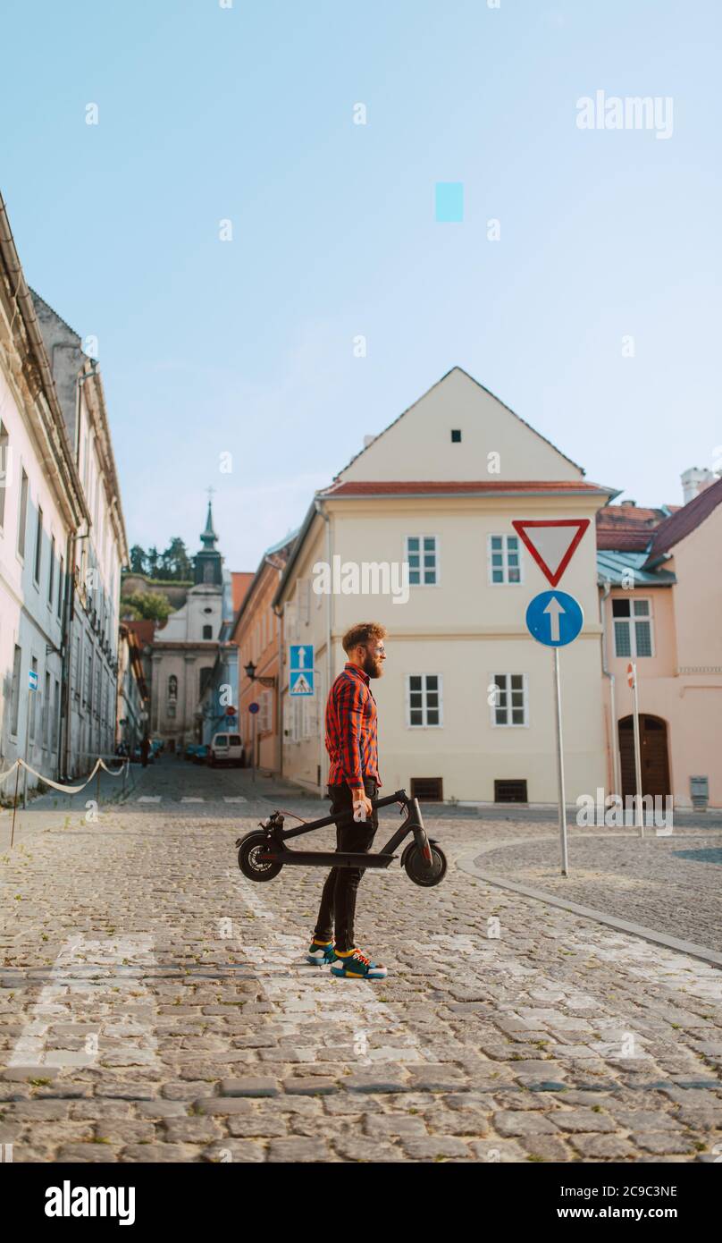 Un beau jeune homme caucasien avec une barbe et des lunettes de soleil se promène au-dessus d'un piéton, tenant un scooter électrique dans la main Banque D'Images