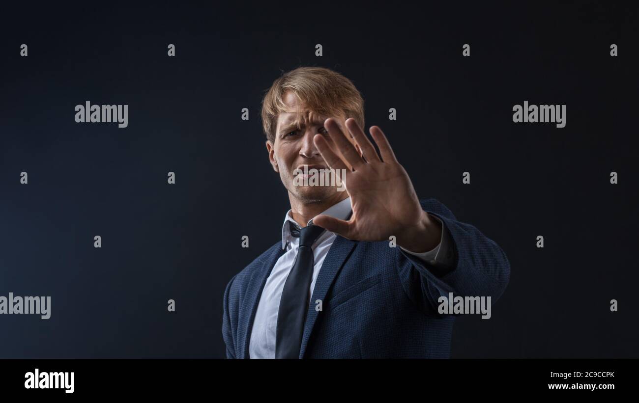 Arrêtez, un jeune homme d'affaires en costume et cravate fait un geste de protestation. Portrait dans le Studio sur un arrière-plan sombre. Banque D'Images
