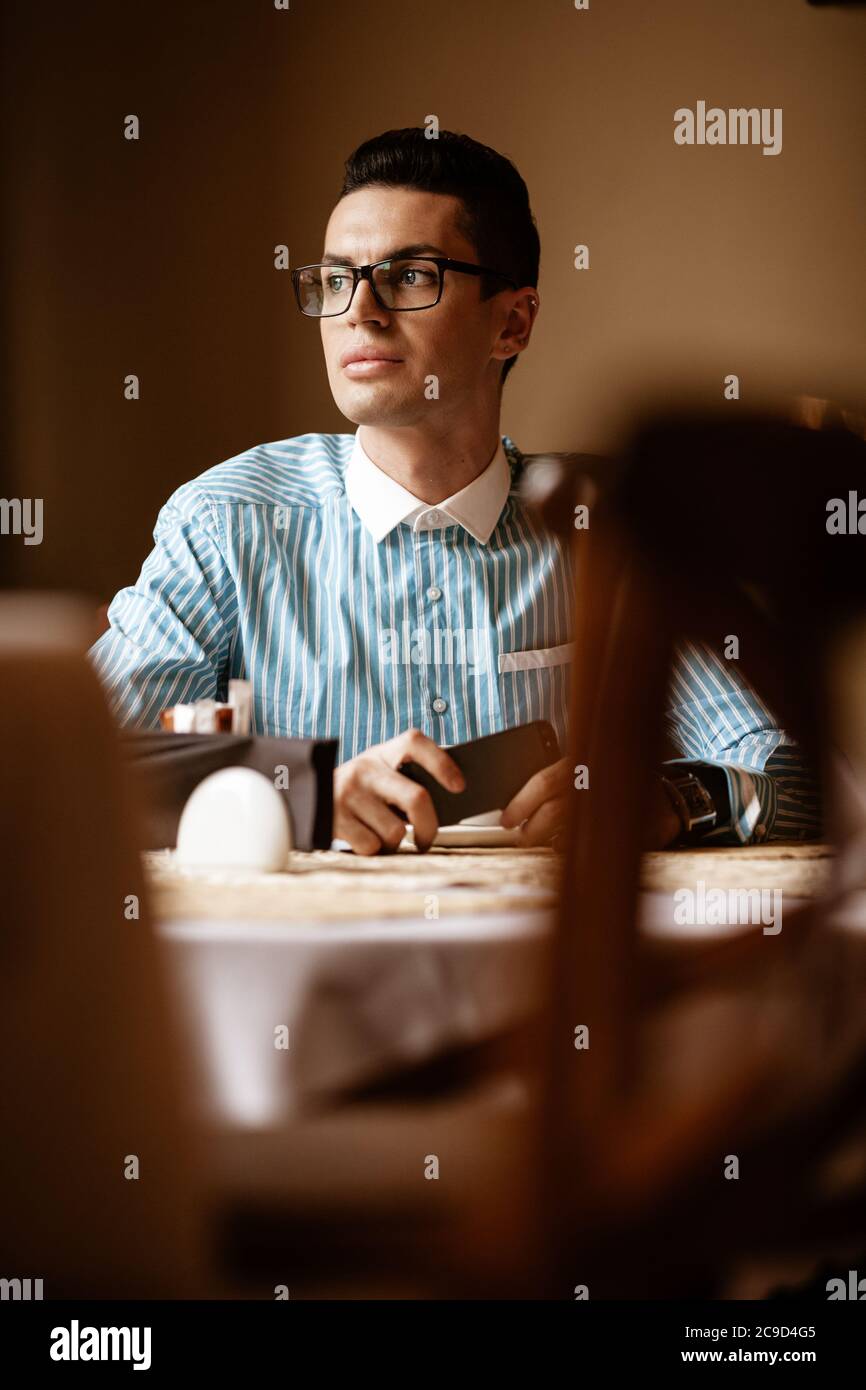 Concept de mode de vie communautaire LGBTQ. Un jeune homosexuel est assis à la table dans un café de la ville à l'ancienne. Beau sourire homme d'affaires gay pose Banque D'Images