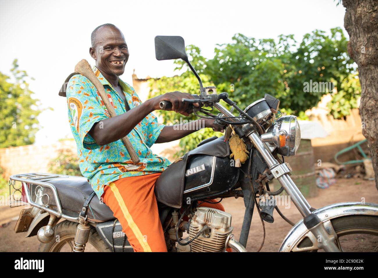 Un fermier mâle chevauche une moto tout en portant une houe de jardinage dans un village rural de la province de Mouhoun, Burkina Faso, Afrique de l'Ouest. Banque D'Images