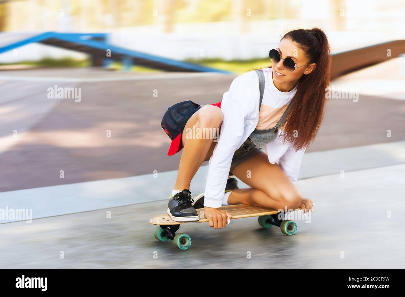 Une fille aux cheveux bruns en lunettes de soleil fait une planche à roulettes dans le parc. Modèle posé sur le terrain de sport Banque D'Images