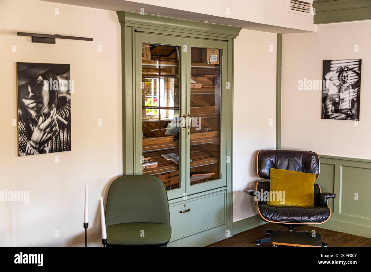 Un cabinet humidor avec des boîtes à cigares se trouve entre deux portraits encadrés en noir et blanc dans l'ancien salon fumeur de l'hôtel Mühle à Binzen, Markgräflerland, Bade-Wurtemberg, Allemagne, avec un fauteuil en cuir et un coussin jaune devant lui. Banque D'Images