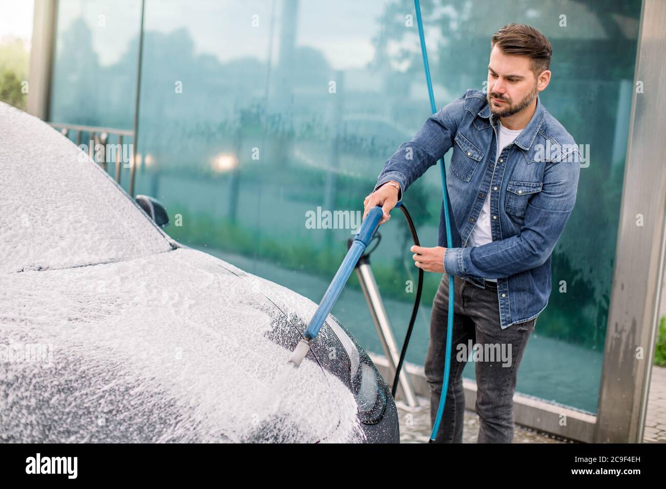 Nettoyage extérieur de la voiture avec de la mousse savonneuse à l'aide d'un jet haute pression. Beau jeune homme barbu lavant sa voiture électrique sous haute pression avec de la mousse Banque D'Images