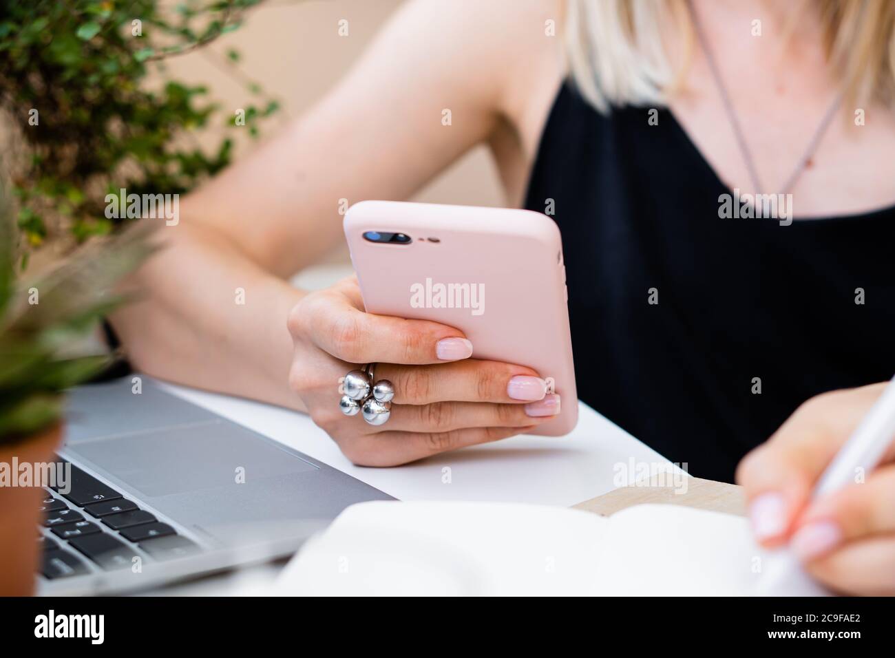 Une femme tient son smartphone à la main et prend des notes dans un bloc-notes dans un bureau confortable d'été avec ordinateur portable Banque D'Images