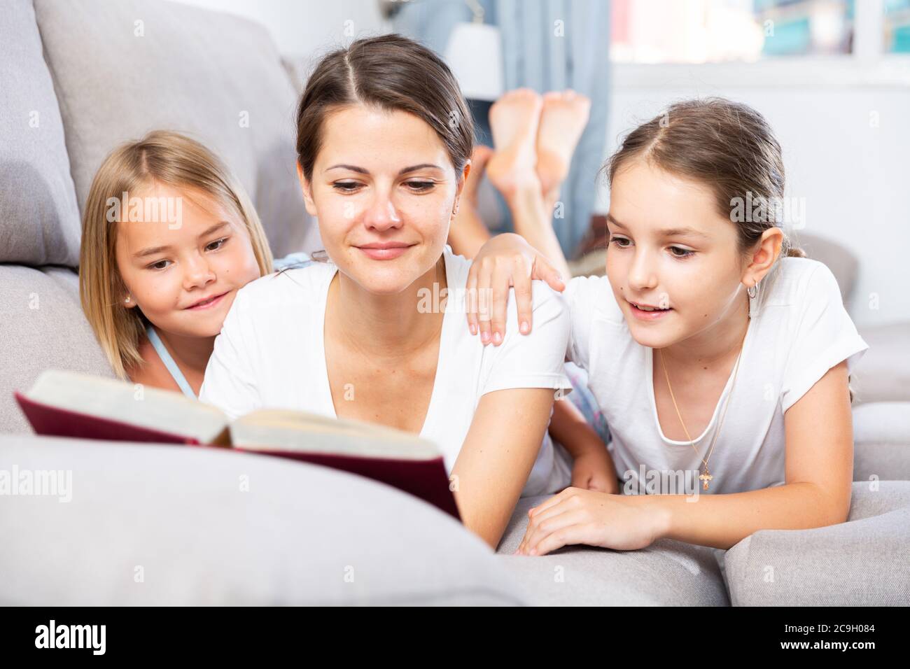 Deux petites filles avec une mère se détendant sur un canapé et lisant un livre Banque D'Images