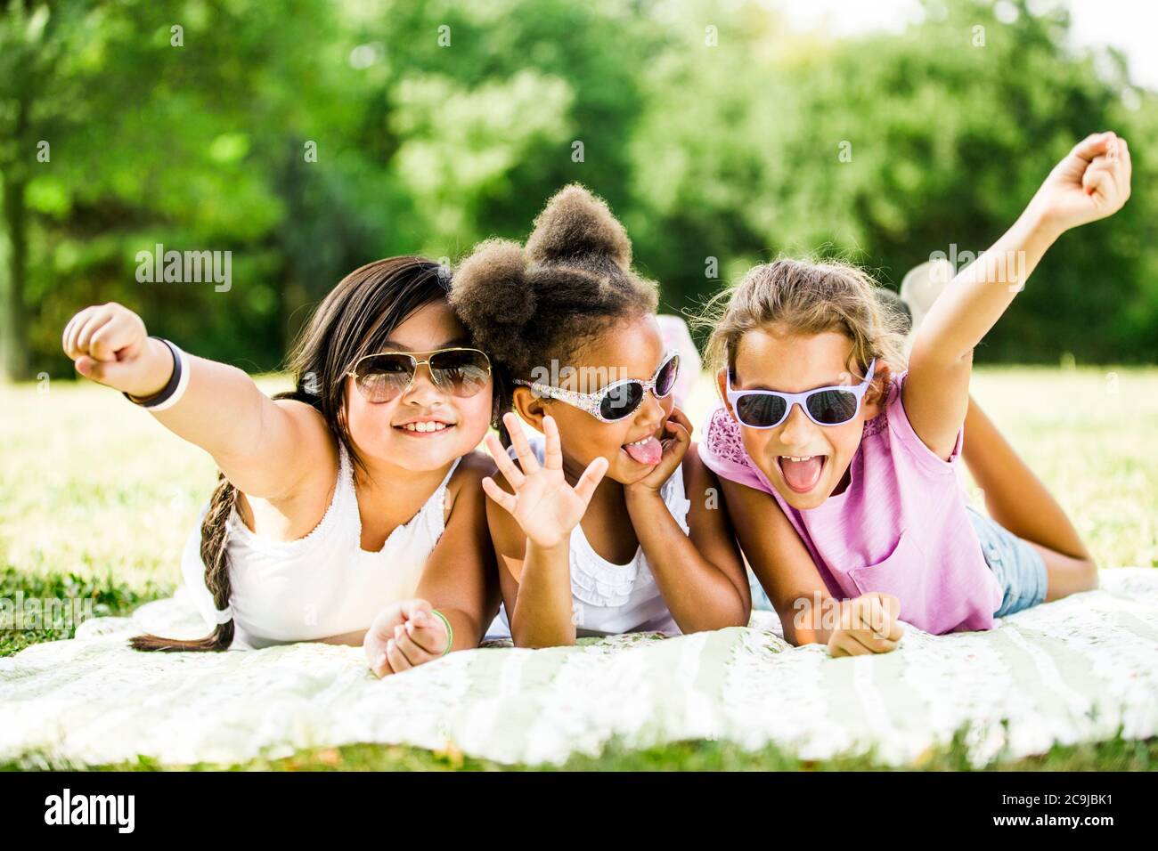 Filles portant des lunettes de soleil et couchée côte à côte sur une couverture dans le parc, souriant, portrait. Banque D'Images