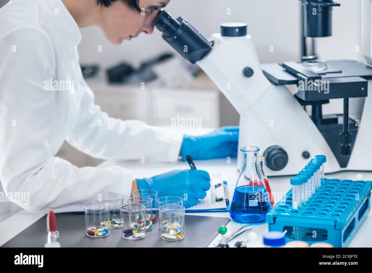 Chercheur médical examinant un nouveau médicament. Femme étudiante en sciences vêtue d'une blouse de laboratoire blanche regardant à travers un microscope. Banque D'Images