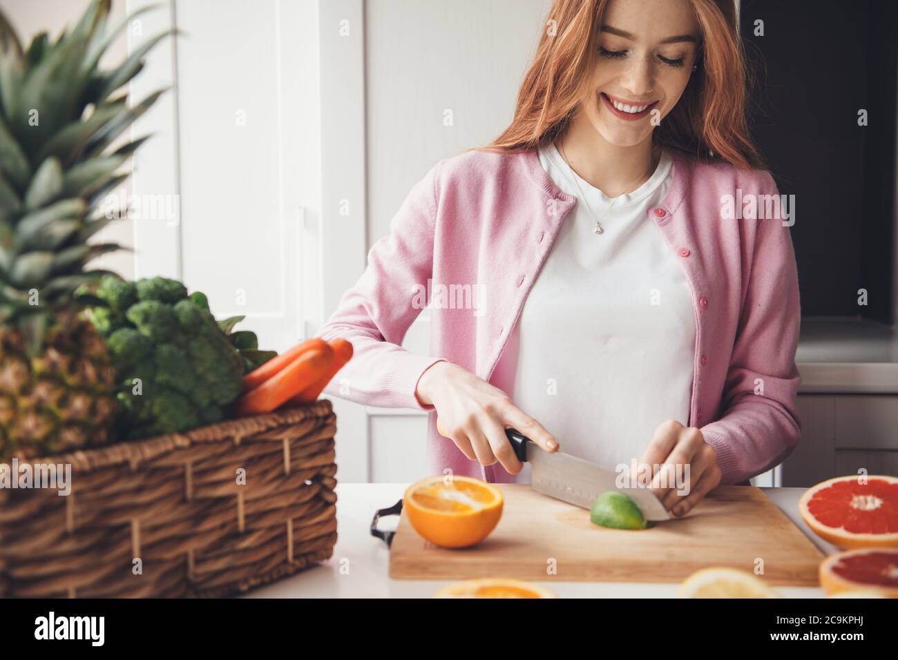 Magnifique femme caucasienne avec des cheveux rouges et des taches de rousseur souriant tout en coupant un citron vert dans la cuisine avec un couteau Banque D'Images