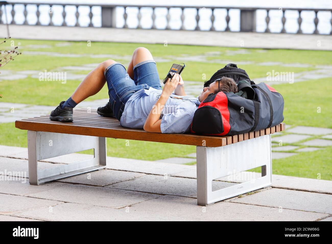 Stockholm, Suède - 31 juillet 2020 : un jeune homme est allongé sur un banc de parc avec un sac comme coussin dans le parc de l'hôtel de ville et utilise son téléphone portable. Banque D'Images