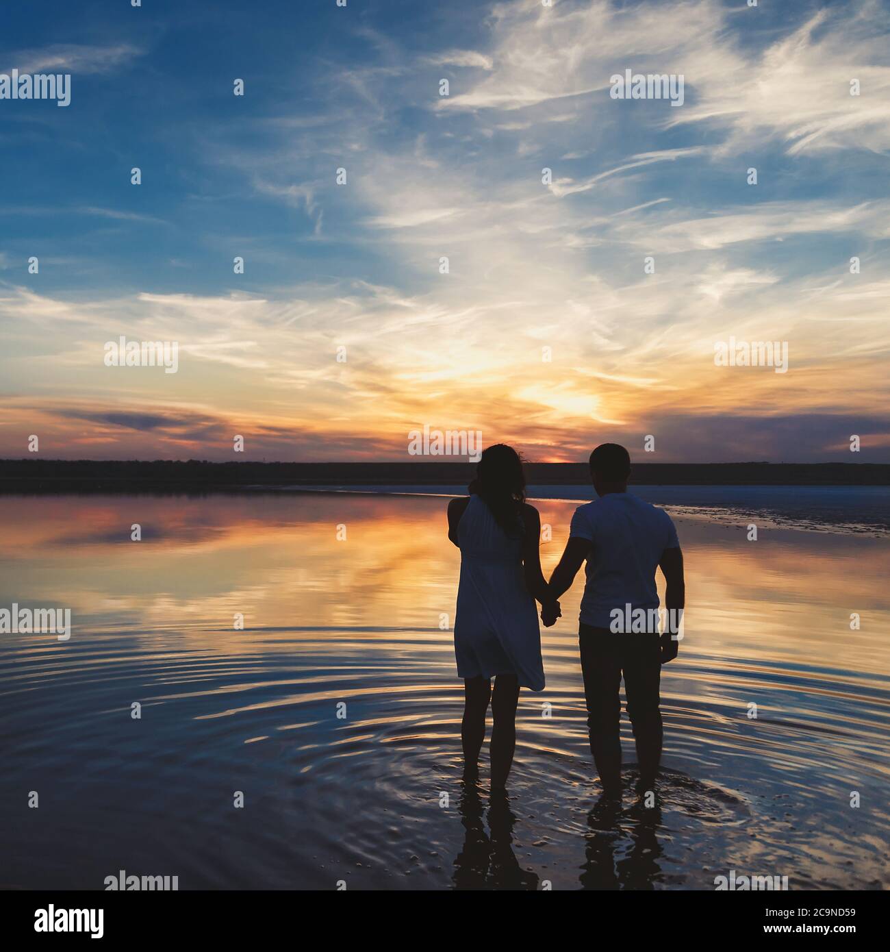 Portrait, silhouette de couple heureux regardant le coucher de soleil coloré lumineux debout dans le grand lac, réflexion dans l'eau, tenant les mains Banque D'Images