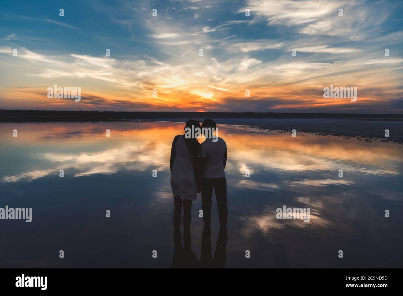 Portrait, silhouette de couple heureux regardant le coucher de soleil coloré et lumineux se tenant dans le grand lac et embrassant, réflexion dans l'eau, tenant les mains Banque D'Images