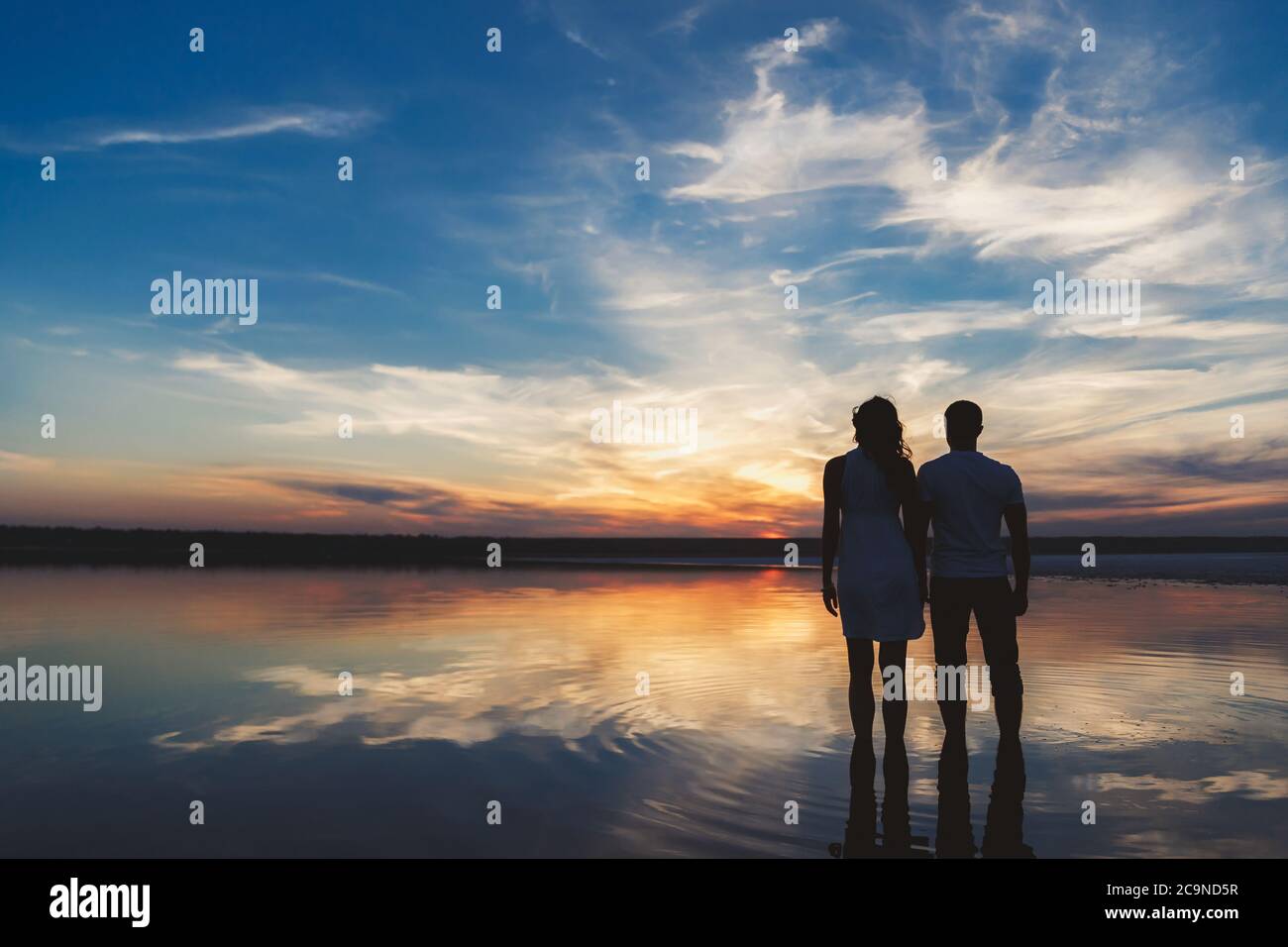 Portrait, silhouette de couple heureux regardant le coucher de soleil coloré lumineux debout dans le grand lac, réflexion dans l'eau, tenant les mains Banque D'Images