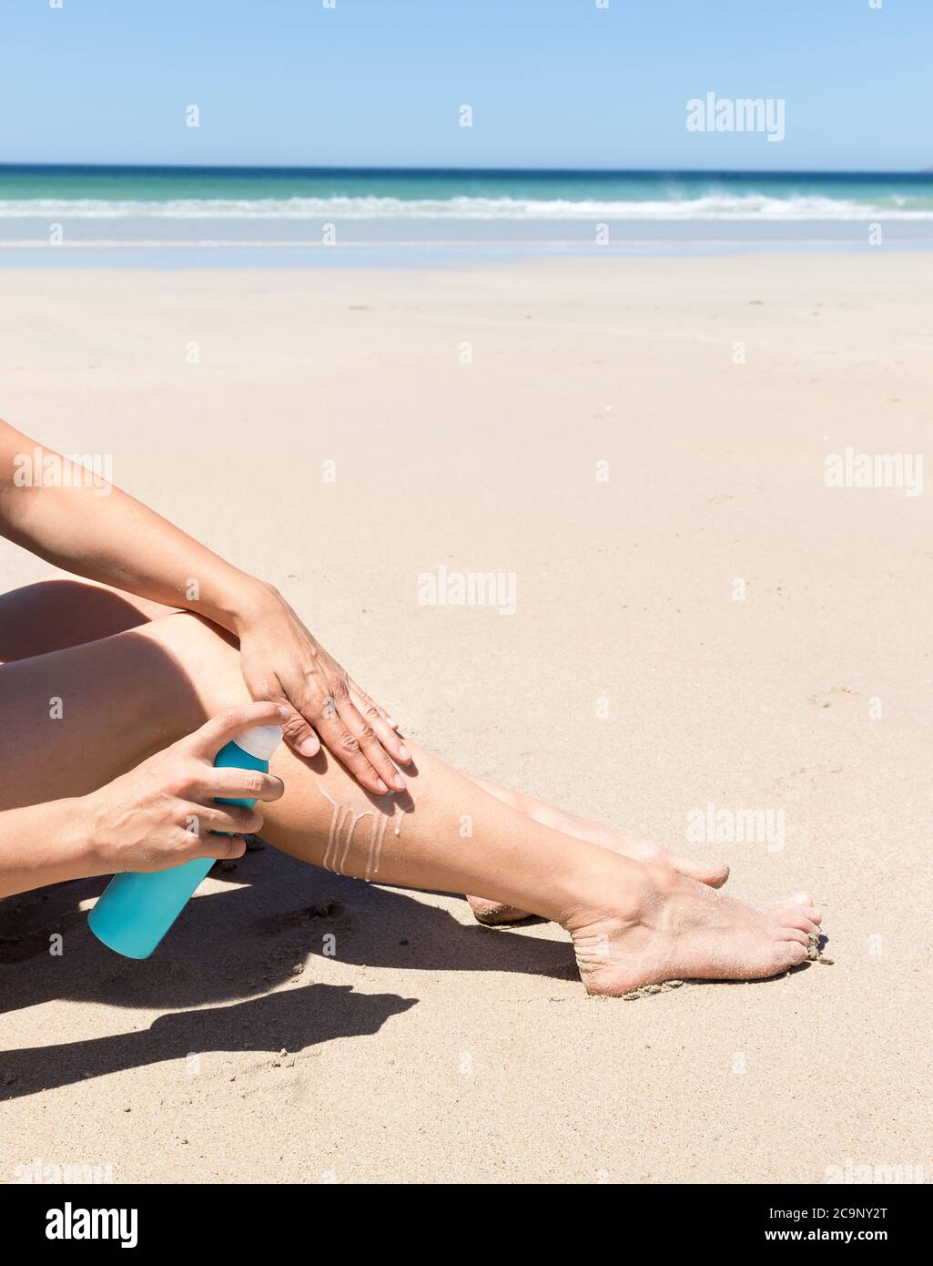 Femme appliquant de la crème solaire sur ses jambes. Assis sur le sable avec la mer en arrière-plan. Banque D'Images