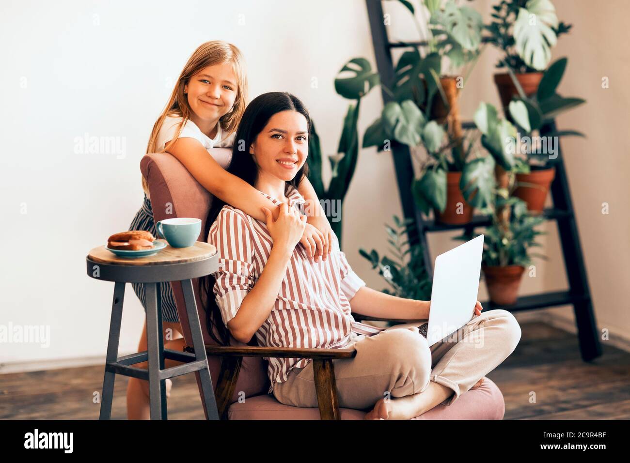 Adorable fille embrassant une jeune femme travaillant sur un ordinateur portable dans un salon confortable et regardant l'appareil photo Banque D'Images