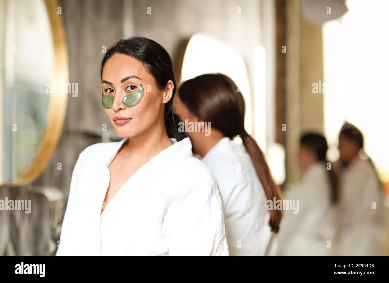 Charmante femme en peignoir et avec des cellules oculaires debout dans la salle de bains moderne tout en faisant soin de la peau quotidien et regardant l'appareil photo Banque D'Images