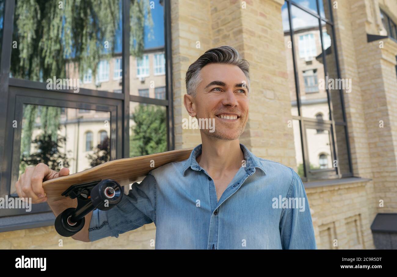 Заголовок Portrait d'un modèle de mode élégant et mature portant des vêtements décontractés, posant pour les photos. Bonne patineuse tenant une longue planche, marchant dans la rue Banque D'Images