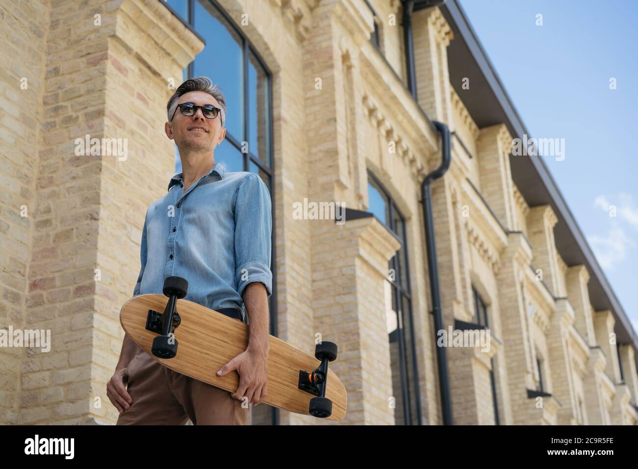 Bonne patineuse tenant une longue planche, marchant dans la rue. Portrait de modèle de mode mature portant des vêtements décontractés et des lunettes de soleil posant pour les photos Banque D'Images