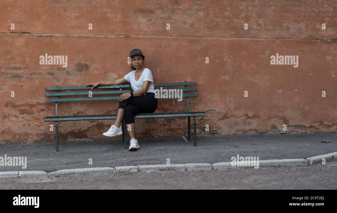 Femme en chapeau, t-shirt, pantalon noir et baskets assis sur un banc de parc avec les jambes croisées à Bologne avec un autre banc de parc à sa droite Banque D'Images
