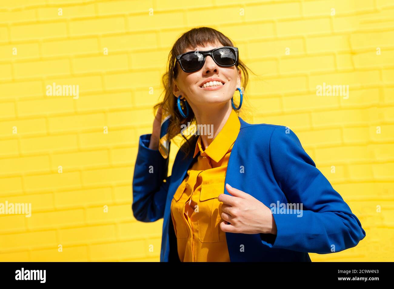 bonne fille sur fond jaune. Une fille en lunettes de soleil, une chemise jaune et une veste bleue se tient contre le mur et sourit. Banque D'Images