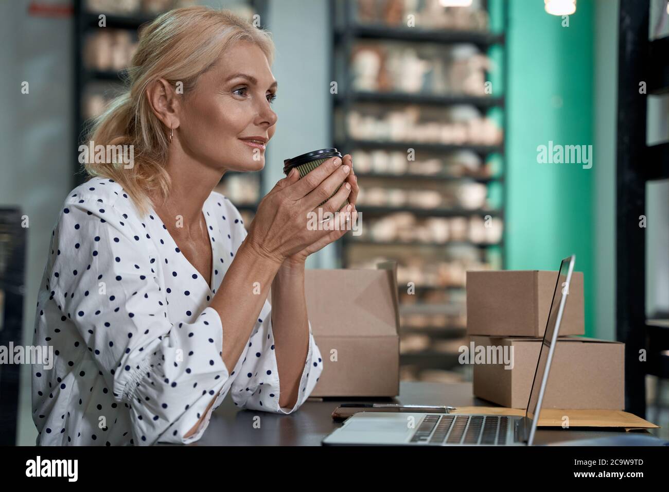 Prendre un café le matin. Vue latérale d'une belle et heureuse femme d'affaires propriétaire ou femme d'affaires buvant un café tout en travaillant dans son studio d'art ou magasin de poterie d'artisanat. Petite entreprise, vente au détail Banque D'Images
