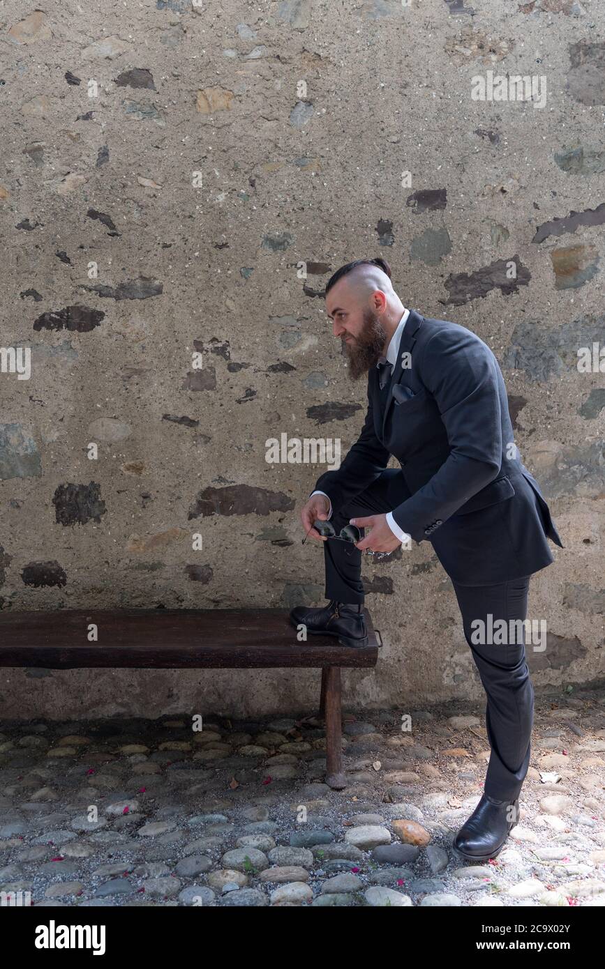 Homme dans des vêtements élégants repose un pied sur un banc en bois dans un petit village Banque D'Images
