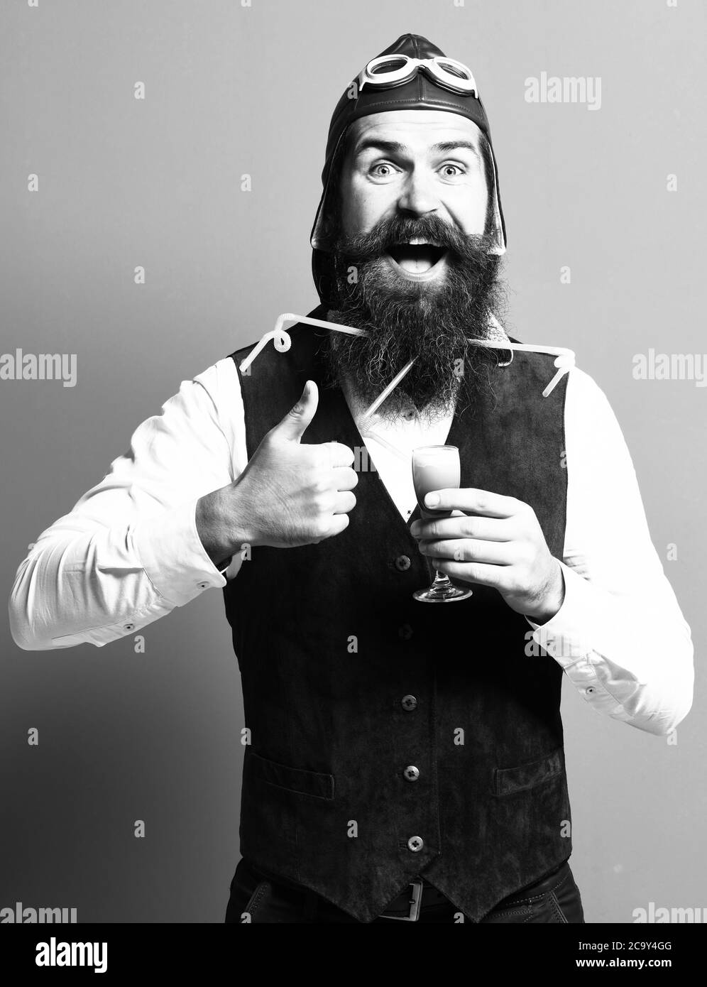 beau pilote barbu ou aviateur avec une longue barbe et une moustache sur le visage souriant tenant un verre de liqueur alcoolique dans un gilet en daim vintage avec chapeau et lunettes, sur fond rouge studio Banque D'Images