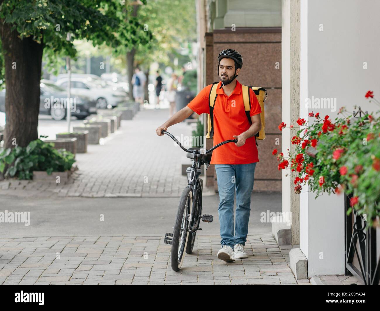 Coursier avec barbe, casque et sac jaune, marche avec vélo dans la rue Banque D'Images