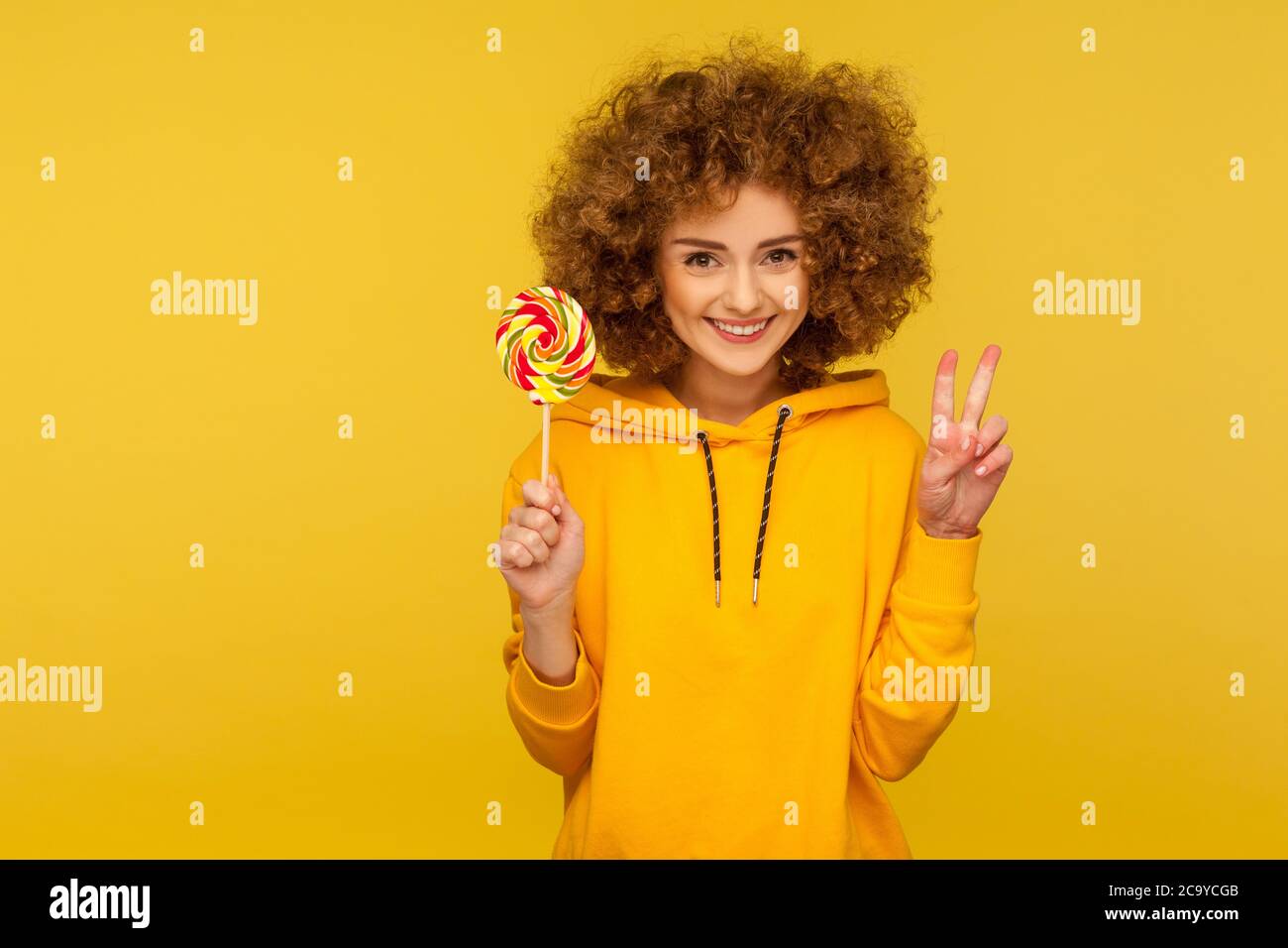 Sucreries arc-en-ciel. Portrait de la femme moderne heureuse à cheveux bouclés en sweat à capuche de style urbain tenant le lopop et montrant le geste de la victoire, souriant à l'appareil photo Banque D'Images