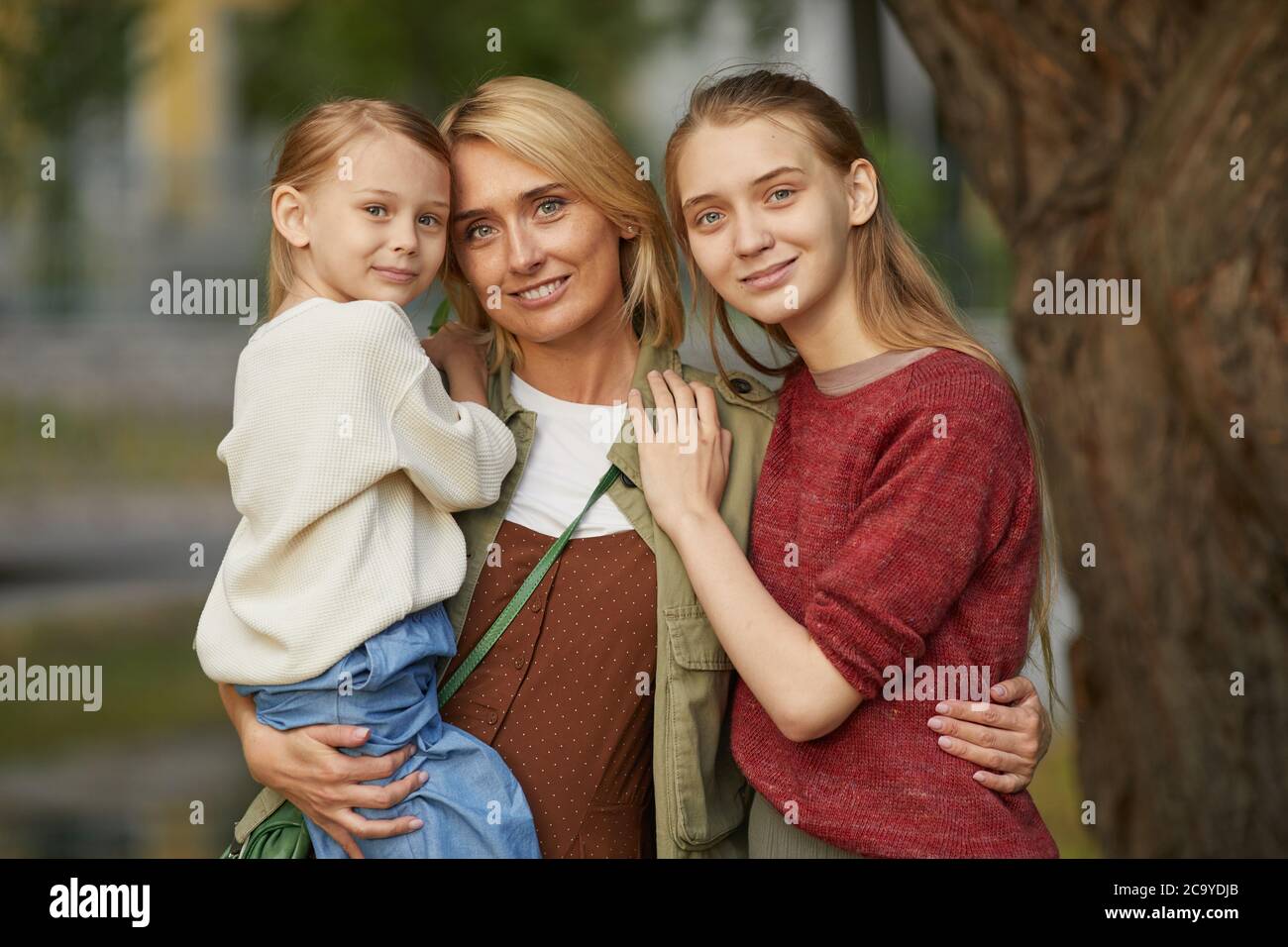 Faites la taille d'une mère adulte moderne avec deux filles qui se posent ensemble et sourient en regardant l'appareil photo tout en vous tenant debout près d'un arbre à l'extérieur, en appréciant le temps passé en famille dans le parc. Banque D'Images